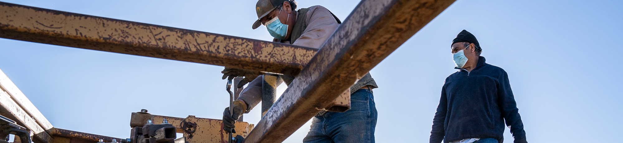 Two farmworkers wearing masks work on a piece of equipment